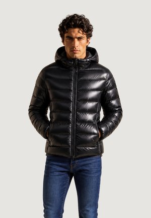 Mand iført en skinnende sort dunjakke med hætte og blå jeans, stående med hænderne i lommerne mod en ensfarvet baggrund.