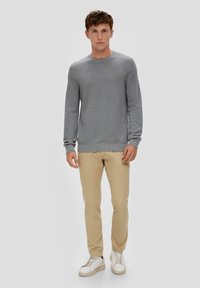 Pull en tricot gris à col rond, avec des poignets et un ourlet côtelés, porté avec un pantalon chino beige et des baskets blanches, se tenant sur un fond neutre.