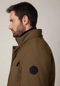 Brązowa ocieplana kurtka z wysokim kołnierzem, zamkiem błyskawicznym, kieszenią na piersi oraz okrągłą naszywką z logo na lewym rękawie. Teksturowany materiał.