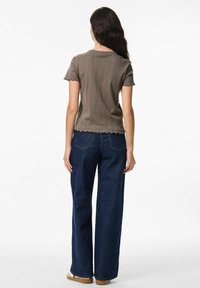 Harmaa kohokuvioitu lyhythihainen paita, jossa on aaltoileva helma, yhdistettynä leveälahkeisiin tummansinisiin farkkuihin. Malli on näkyvissä selkäpuolelta tasaisella taustalla.