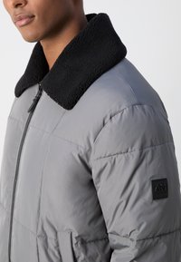 Doudoune grise avec un col noir en peluche, design matelassé, fermeture éclair à l'avant et un patch logo sur la manche gauche. Texture lisse.