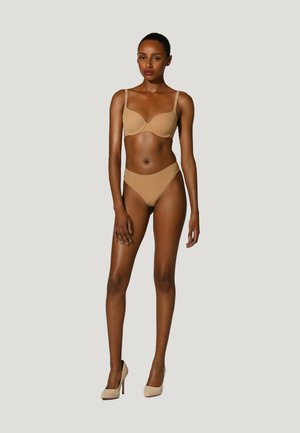 Kvinde stående i beige bh, matchende undertøj og beige højhælede sko mod en ensartet lys baggrund.