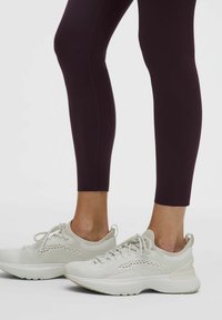 Lengvi sportiniai batai kremine spalva, su orui pralaidžiu tinklinio audinio viršumi su perforacijomis, derinami su tamsiai violetinėmis aptemptomis tamprėmis.