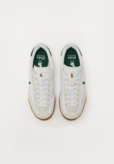 Sapatilhas brancas com detalhes verdes, apresentando um logo dourado, materiais em camurça e couro, sola de borracha e design de atacadores.