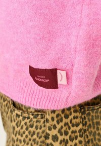 Ružový pletený sveter má rebrovaný lem a hnedú textilnú nálepku s názvom značky "NAOKO Knitwear." Nosený cez nohavice s leopardím vzorom.