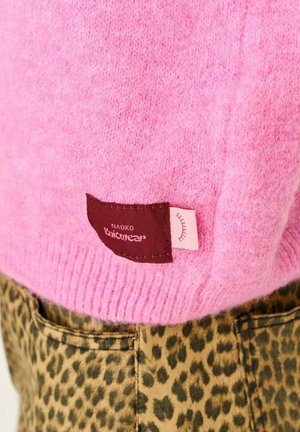 Różowy wełniany sweter z ribbowym wykończeniem i brązową metką z nazwą marki "NAOKO Knitwear". Nosi się go z spodniami w panterkę.