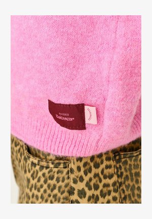 Różowy wełniany sweter z ribbowym wykończeniem i brązową metką z nazwą marki "NAOKO Knitwear". Nosi się go z spodniami w panterkę.