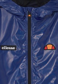 Niebieska błyszcząca kurtka z czarnym zamkiem, naszywką z nazwą marki "ellesse" na lewym piersi oraz pomarańczowo-czerwonym logo w kształcie półksiężyca na prawej piersi.