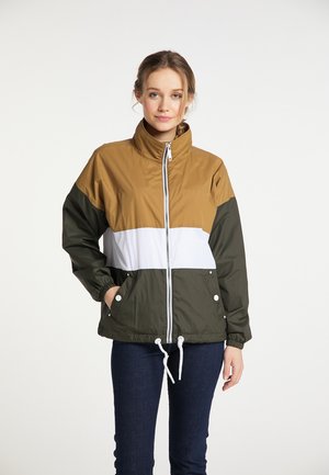 Frau trägt eine farbblockierte Windjacke in Braun, Weiß und Dunkelgrün, steht mit einer Hand in der Tasche vor weißem Hintergrund.