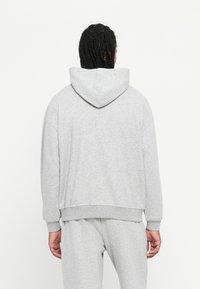 Osoba s pletenicama koja nosi svijetlosivu duksericu i odgovarajuće trenirke, stoji okrenuta leđima prema jednostavnoj bijeloj pozadini.