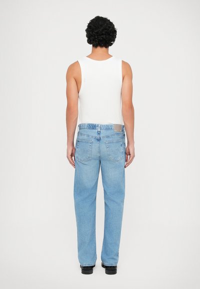 Heledad sinine denimist teksapüksid, millel on mugav lõige, koos valge ilma varrukatetopiga. Omadusteks on tagakotid ja nahkpael vööl.