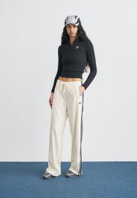 Γυναίκα που φοράει μαύρο κοντό τοπ με φερμουάρ, εκρού φόρμα με ναυτικά ρίγες, ασημένια παπούτσια και λευκό-μαύρο καπέλο, στέκεται σε μπλε χαλί.