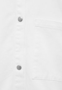 Camicia bianca in cotone con una texture liscia, dotata di bottoni a pressione argentati e una tasca quadrata sul petto. Design semplice.