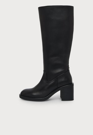 Botte noire en cuir montée jusqu’au genou avec bout arrondi et talon bloc épais, montrée de l’intérieur sur fond blanc.