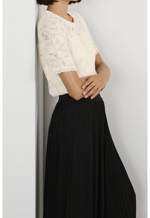 Femme appuyée contre un mur portant un top en dentelle crème à manches courtes et une jupe plissée noire taille haute, bras croisés.