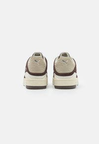 Beige ja pruuni Puma tossud, nähtud tagant, logoga kandadel ja kreemivärvi mööbliga valgel taustal.