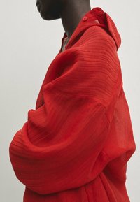 Chemise rouge texturée à coupe décontractée, avec des détails à boutons et une capuche. Le tissu paraît léger et semi-transparent, mettant en valeur de fines lignes horizontales.