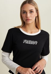 T-shirt nera a maniche corte sovrapposta a una maglietta bianca a maniche lunghe. Presenta il logo "FREDDY" in grassetto con testo a righe bianche nella parte frontale.