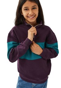 Leende tjej i en lila och teal zip-up tröja, håller dragkedjan med båda händerna, klädd i blå jeans.