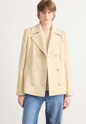 Light beige double-breasted wool coat with a notched collar, visible buttons, and side pockets. Worn over a beige sweater and blue jeans.