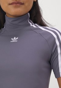 Šedý fitted top Adidas s vysokým límcem, který má tři bílé pruhy na ramenou a malé bílé logo na hrudi.