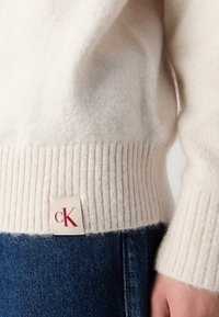 Kremfarget strikket genser med ribbestrikkede mansjetter og kant, med en merkelapp som viser "cK." Båret over blå denimjeans, som viser teksturkontraster.
