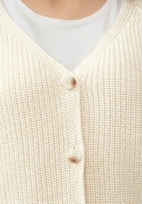 Strickjacke in creme mit V-Ausschnitt, ausgestattet mit zwei runden Knöpfen und einem strukturierten Stichdetail. Darunter ist ein weißes Hemd sichtbar.