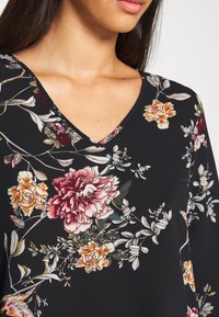 Femme portant un haut noir à col en V avec un imprimé floral détaillé dans des tons de rose, orange, blanc et vert, montrant l'encolure et le haut de la poitrine.