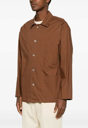 Uomo con una camicia marrone abbottonata con tre tasche frontali, abbinata a pantaloni beige chiaro, in piedi davanti a uno sfondo neutro.