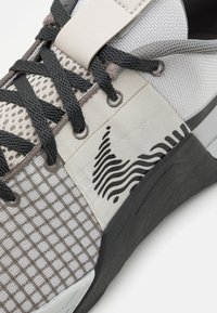 Szaro-biały but sportowy z teksturowaną siateczką, wzorem w kratkę, czarną gumową podeszwą i charakterystycznym logo fali po boku.