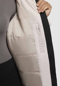Main tenant tenant d'une veste matelassée beige ouverte montrant la poche intérieure, portée sur un haut gris et un bas noir.