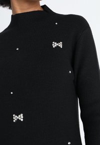 Pull noir à côtes avec de petites accents décoratifs en cristal, incluant un motif de nœud. Encolure haute avec une coupe ajustée.