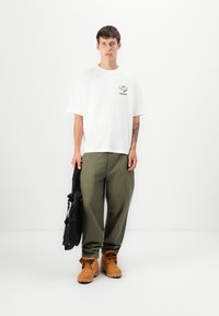 White oversized t-shirt with a small printed logo, olive green loose-fitting pants, and tan work boots, holding a black backpack.