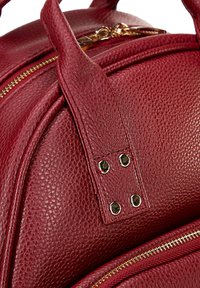 Zaino in pelle rossa dalla texture morbida con dettagli dorati, dotato di chiusura con cerniera e accenti ribattuti sulle cinghie del manico.