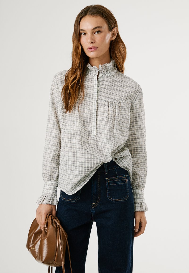 Vit och grå rutad blus med volangkrage och volanger vid ärmarna, ihopparad med mörkblå denimjeans. Modellen håller en brun läderväska.