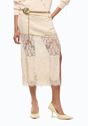 Donna che indossa una gonna midi in pizzo color crema con motivi floreali, una cintura a catena dorata con dettaglio floreale e scarpe con tacco crema.