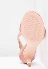 Sandale à talon haut en daim rose unique montrée de la semelle avec un motif ondulé sur une surface et un mur blancs.