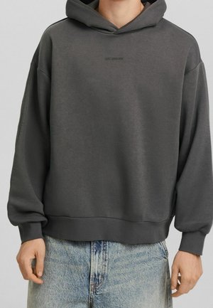 Ciemnoszara oversize bluza z kapturem z małym czarnym napisem na piersi, noszona z jasnoniebieskimi wyblakłymi dżinsami, pokazująca górną część ciała i ręce.
