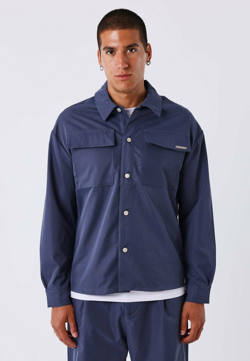 Marineblauwe jas met lange mouwen, voorzien van een klassieke kraag, twee borstzakken en een knoopsluiting aan de voorkant, gemaakt van een gladde stof.