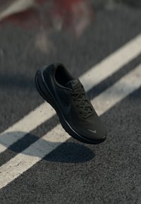 Svart Nike träningssko med ovandel i mesh, rund tå och texturerad sula, svävande över en texturerad yta, utan synliga snören.
