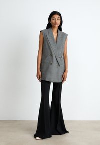 Harmaa raidallinen hihaton bleiseri, jossa on kaksiriviset napit, yhdistettynä mustiin levennettyihin housuihin ja beigecolorisiin korkokenkiin neutraalilla taustalla.