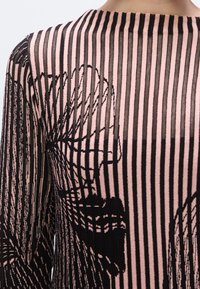 Haut en maille rayé noir et rose avec une texture transparente et un motif floral, encolure ronde et manches longues. Variations subtiles de texture présentes.