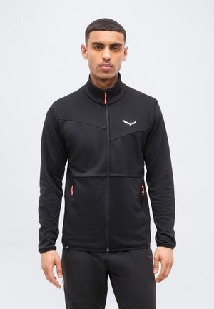 Άνδρας που φοράει μαύρο μπουφάν με φερμουάρ, υψηλό γιακά και φερμουάρ στις πλαϊνές τσέπες, μπροστά σε απλό φωτεινό φόντο.