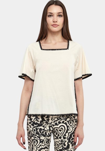 Cream short-sleeve top with a square neckline and black scalloped trim. Paired with black and white patterned pants featuring abstract designs.