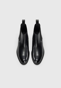 Botas de piel negras hasta el tobillo con textura suave, paneles laterales elásticos, punta redonda y tiradores en la parte trasera. Diseño minimalista.