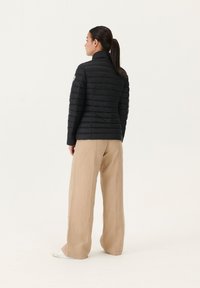 Giacca a vento nera con alto colletto, design a righe verticali e dettaglio logo sulla spalla. Abbinata a pantaloni beige leggeri a gamba larga.