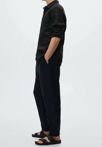 Camisa negra de botones con mangas remangadas, pantalones negros holgados y sandalias negras con tiras, situados sobre un fondo liso.