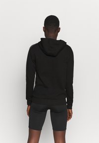 Sweatshirt noir avec capuche, poignets et ourlet côtelés. Le tissu semble doux, présenté de dos avec un short noir assorti.