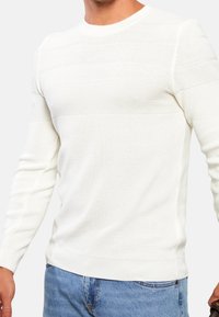 Weißer Strickpullover mit strukturierten horizontalen Streifen, rundem Ausschnitt, langen Ärmeln sowie gerippten Bündchen und Saum. Kombiniert mit blauen Jeans.
