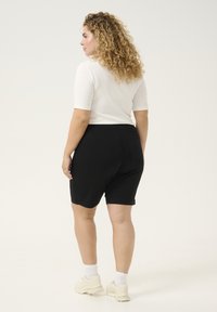 Pantaloni corti neri in cotone misto con una texture liscia, di lunghezza media fino alla coscia, e una tasca posteriore singola. Abbinati a una maglietta bianca aderente a maniche corte.
